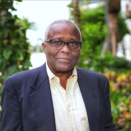 Older Black man with glasses, wearing a blue blazer and yellow shirt, outdoors in a garden.