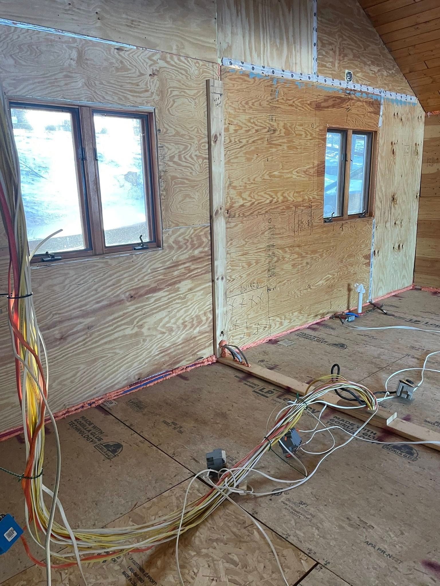 Interior of a building under construction, showing plywood walls, windows, and exposed wiring on the floor.