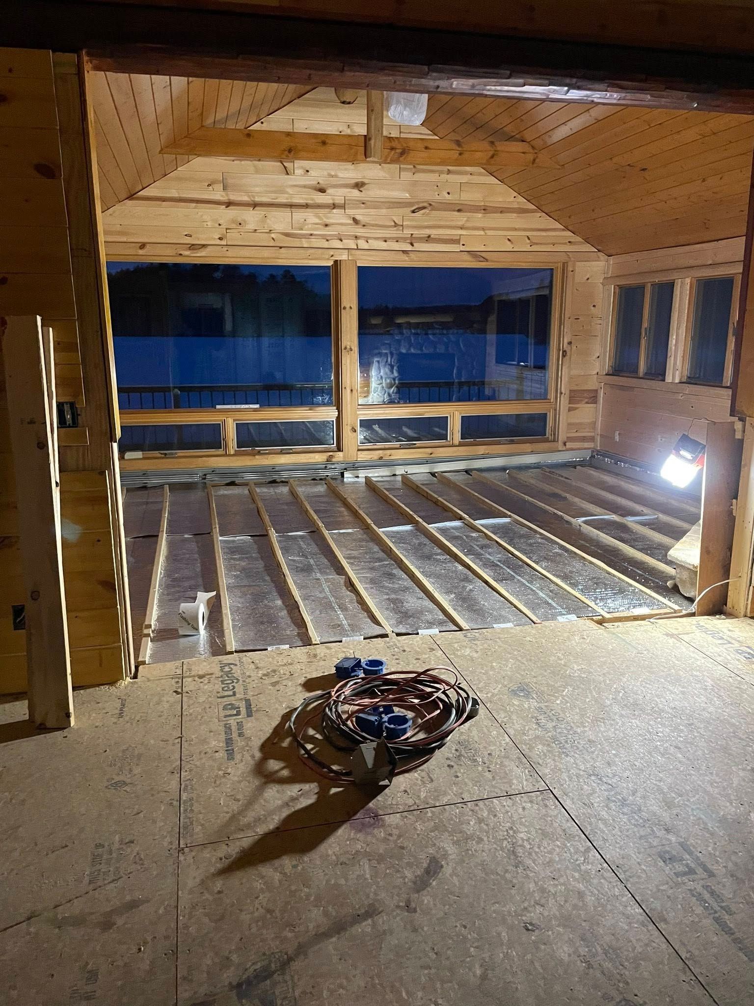 Interior of a wooden room under construction; exposed beams and subfloor, large window, tools on the floor.