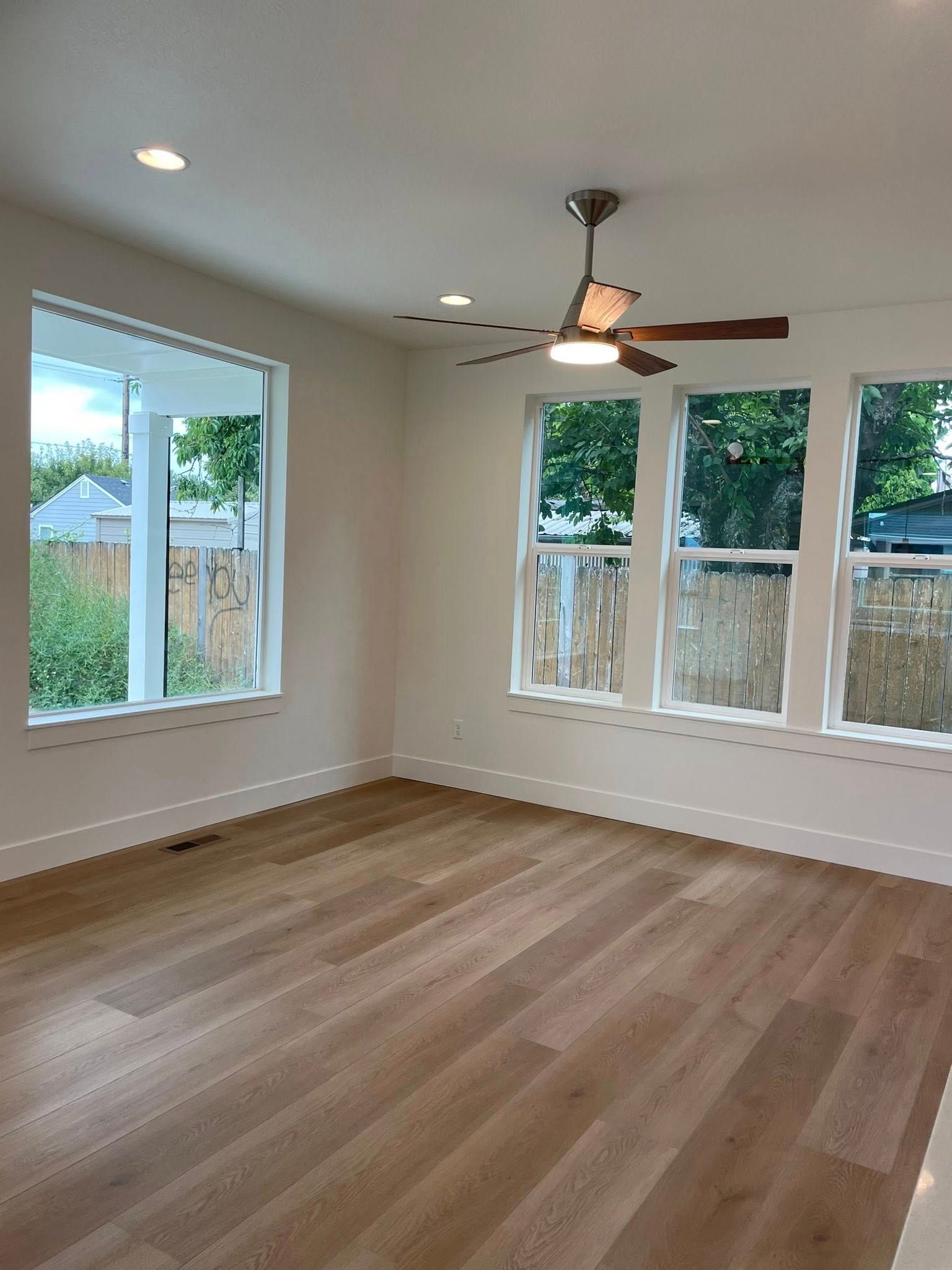 Empty room with hardwood floors, white walls, large windows, and a ceiling fan.
