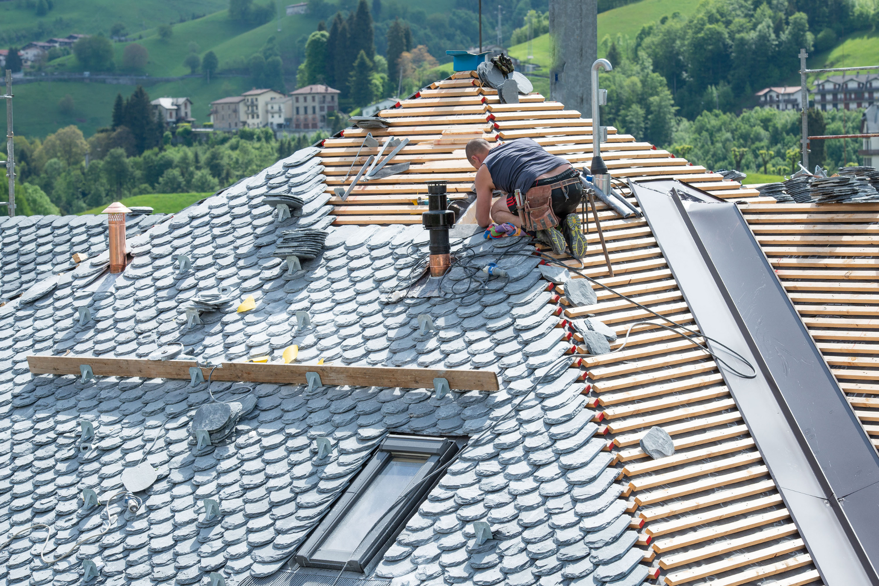 Roofer working on a gray tiled roof with a skylight, with a green, mountainous backdrop.