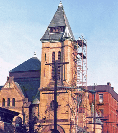 Brick church tower with scaffolding, phone poles, and a red building against a blue sky.