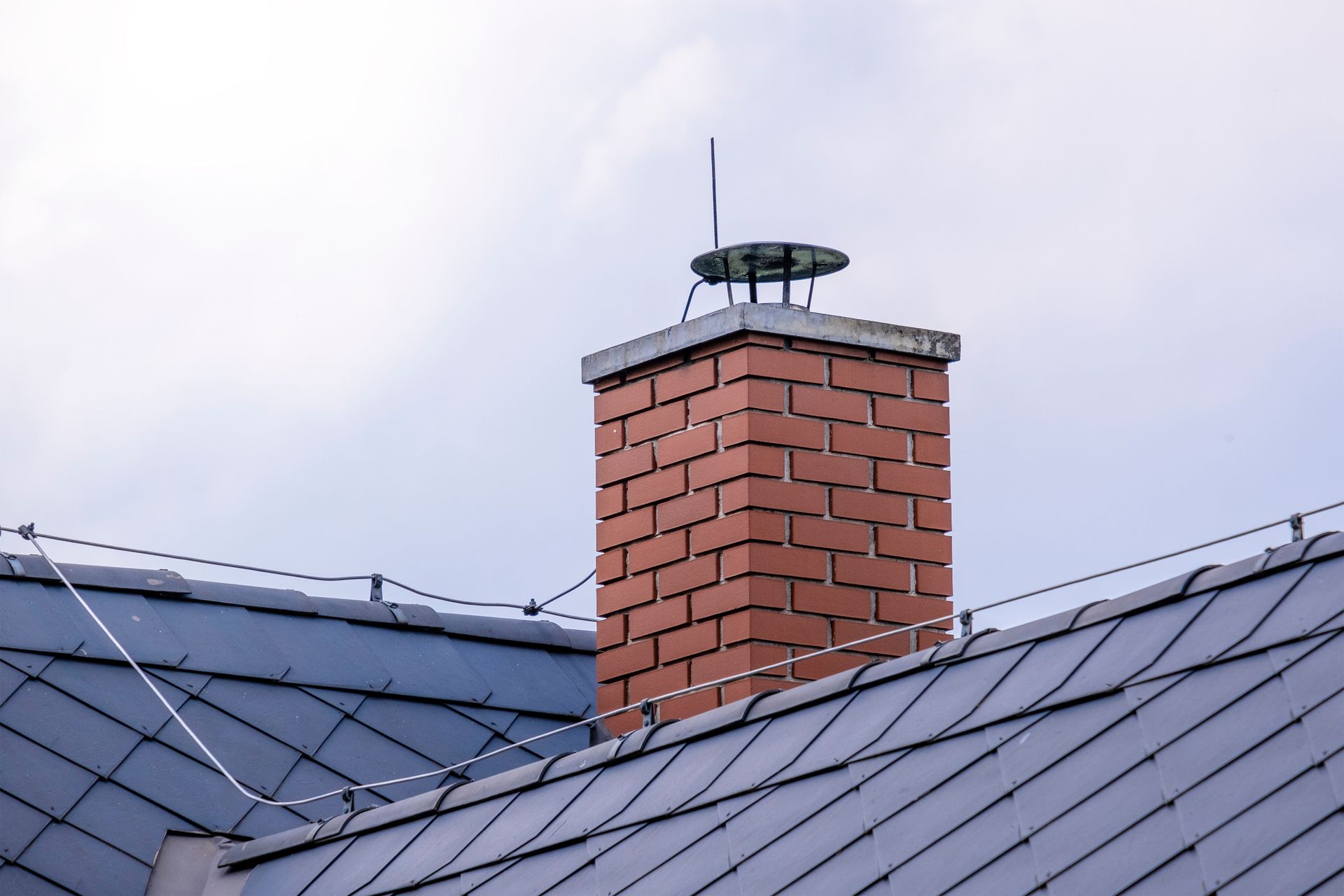 Brick chimney on a gray roof, with a metal cap and antenna against a cloudy sky.