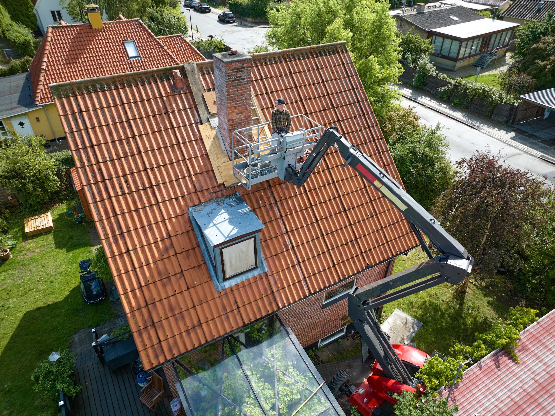 Red-tiled roof with a person in a lift working near a chimney, surrounded by green foliage, blue sky.