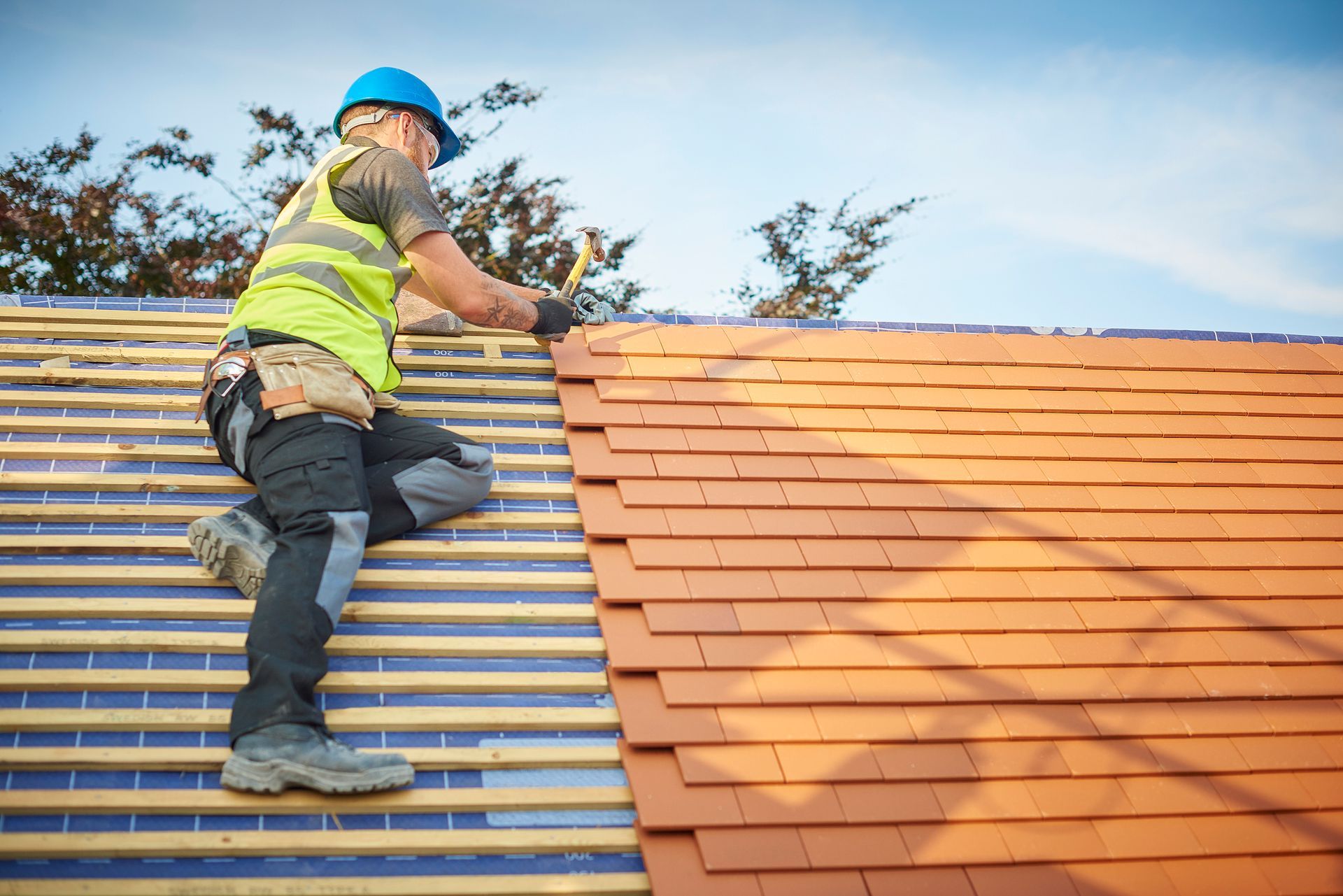 Roofer in safety gear installing clay tiles on a roof. Blue sky in background.