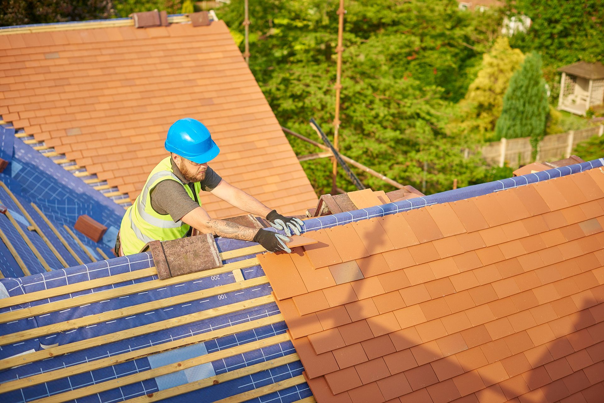 Roofer in safety gear installing clay tiles on a house roof.
