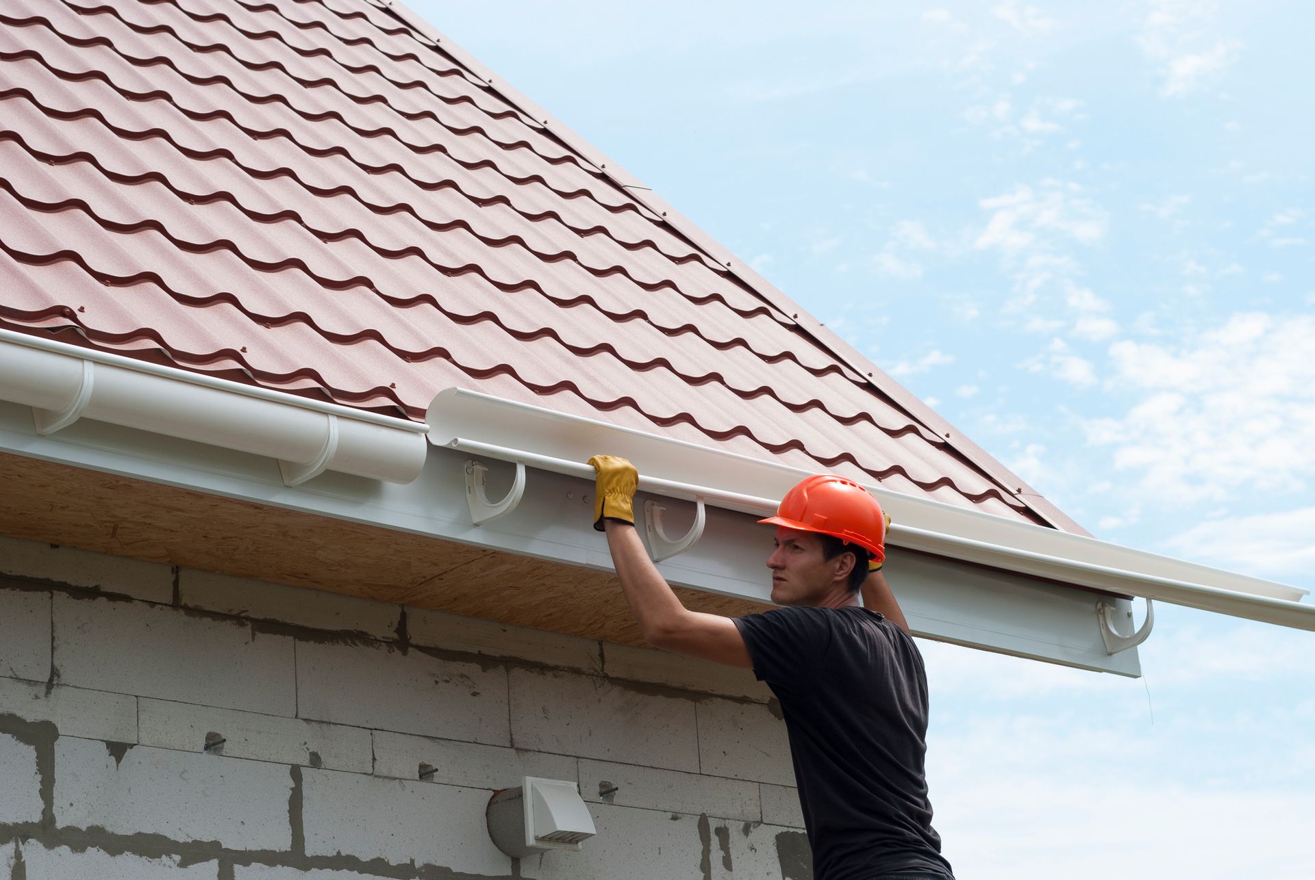 Man in hard hat installing gutter on brick building under red roof.