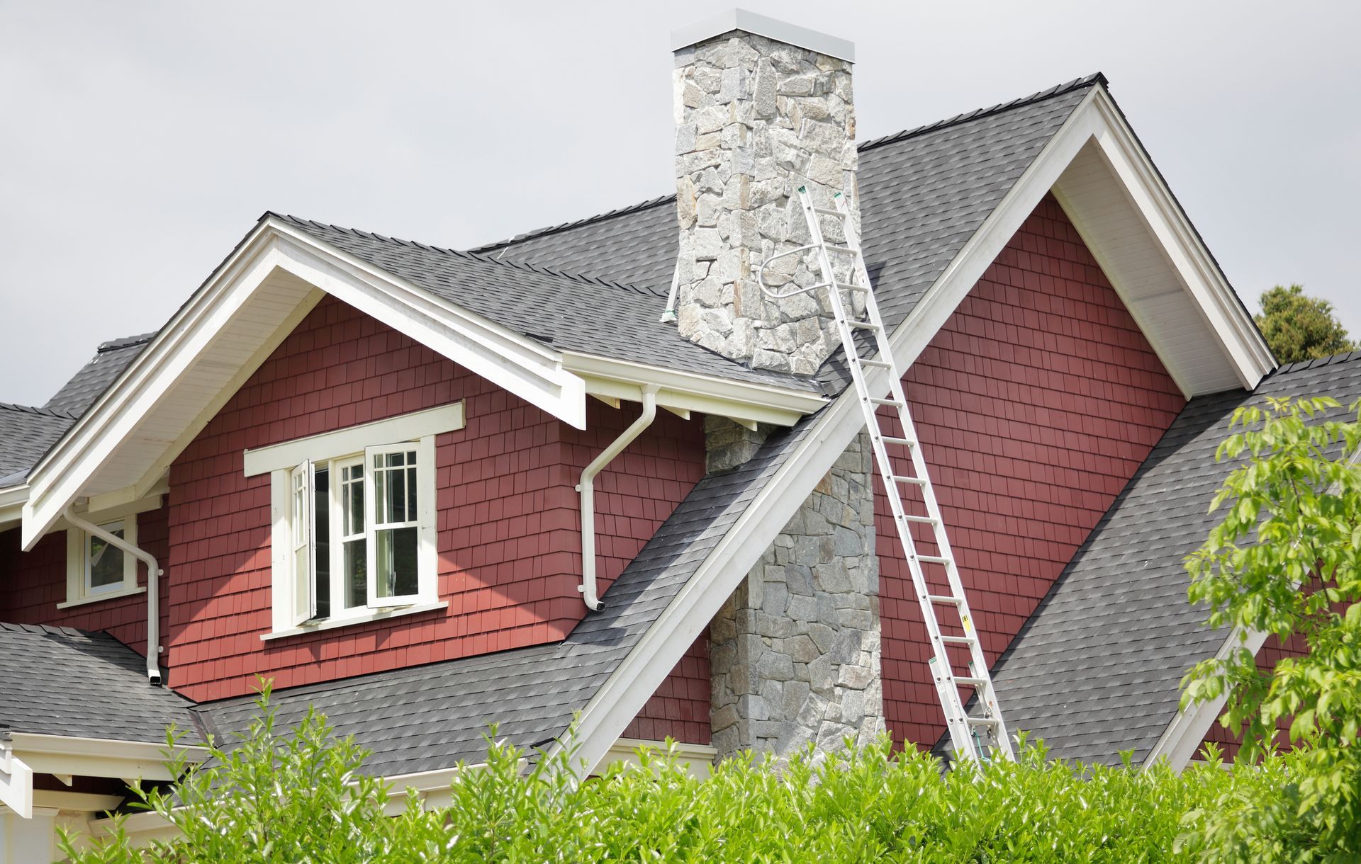 Red house with gray roof, white trim, stone chimney, and ladder leaning against it.