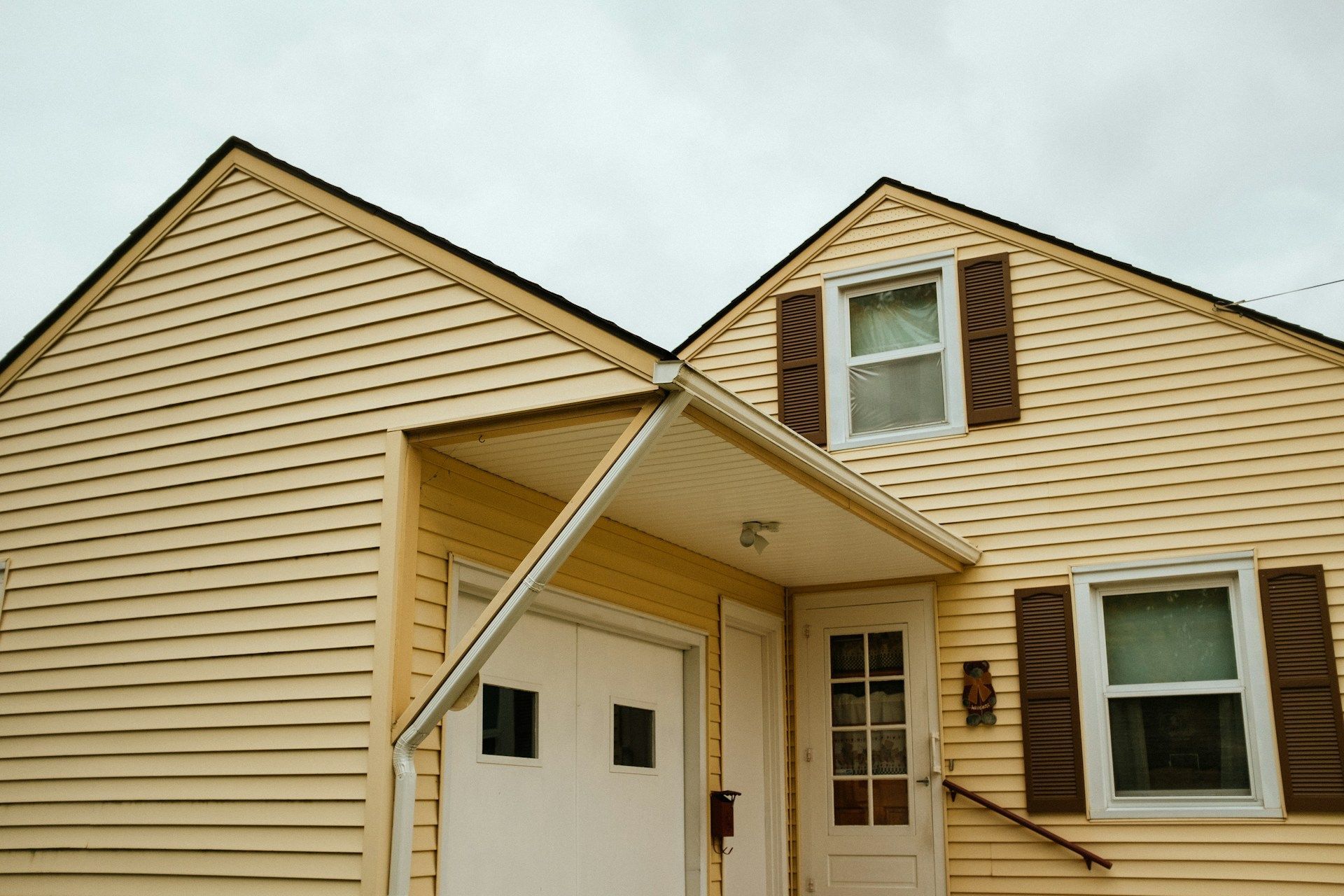 Yellow house with white door and garage door, brown shutters, and cloudy sky.
