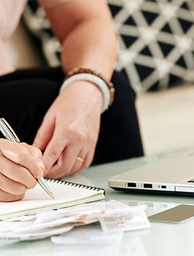 Person writing in a notebook next to a laptop and bills on a table.