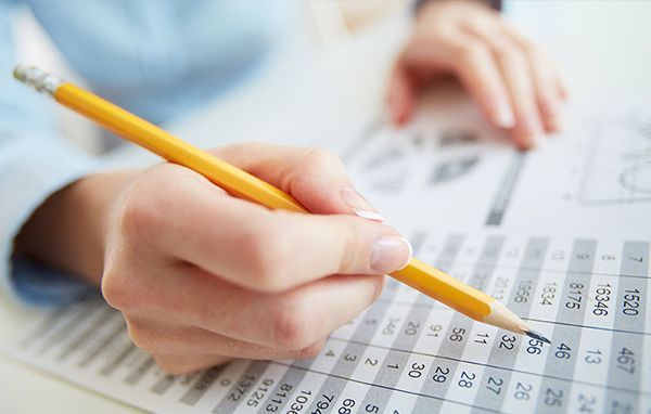 Person's hand holding a pencil, writing or calculating on a spreadsheet with numbers.
