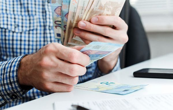 Person counting a stack of Euro banknotes at a desk, wearing a blue plaid shirt.