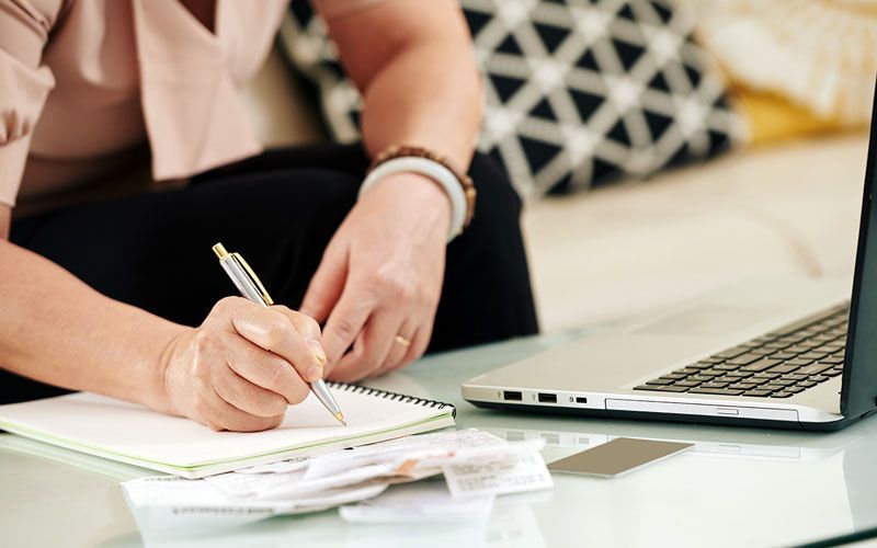 Person writing in a notebook next to a laptop and bills on a table.