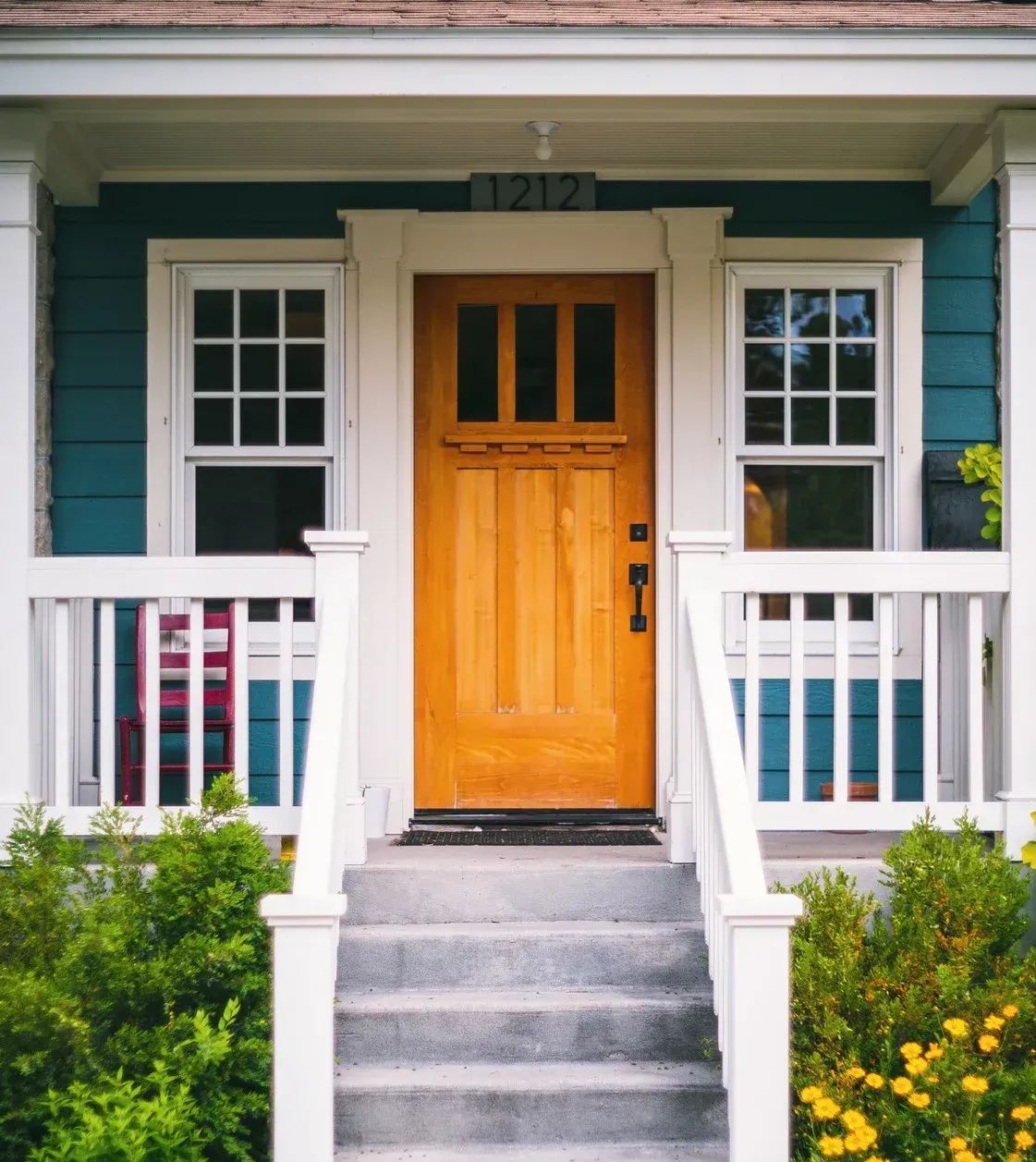 A front porch with a wooden door, teal siding, white railings, and steps leading up to the entrance.