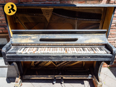 Old, weathered piano outdoors, keys visible, black exterior, top open. Brick wall background.