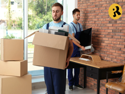 Movers carrying boxes and a computer monitor inside a room with a desk.