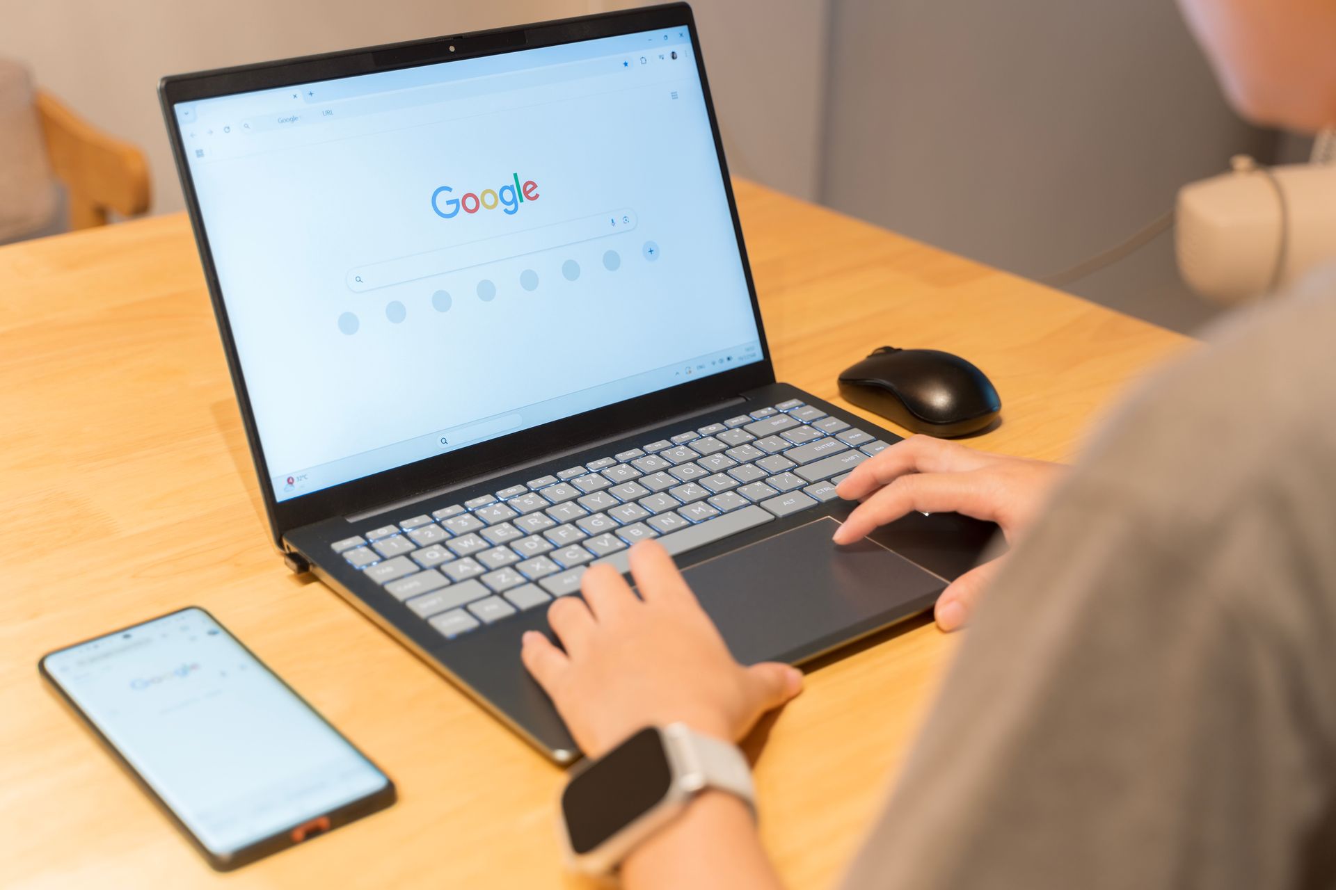 Person using a laptop with Google search open, a mouse, and a smartphone on a wooden table. Person using a laptop with Google search open, a mouse, and a smartphone on a wooden table.