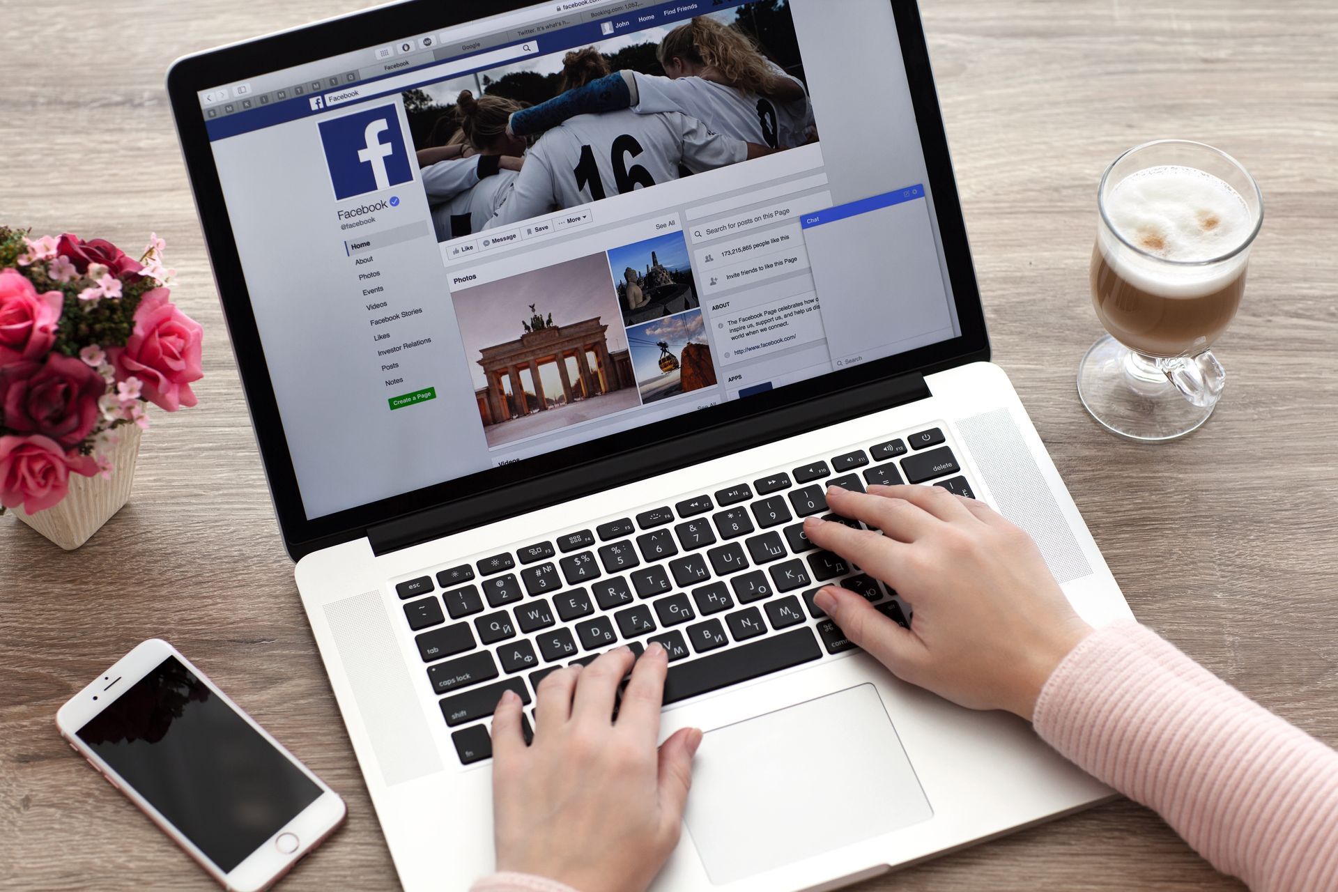 Person using a laptop with Facebook open, typing on the keyboard. A phone, flowers, and a drink sit nearby. Person using a laptop with Facebook open, typing on the keyboard. A phone, flowers, and a drink sit nearby.