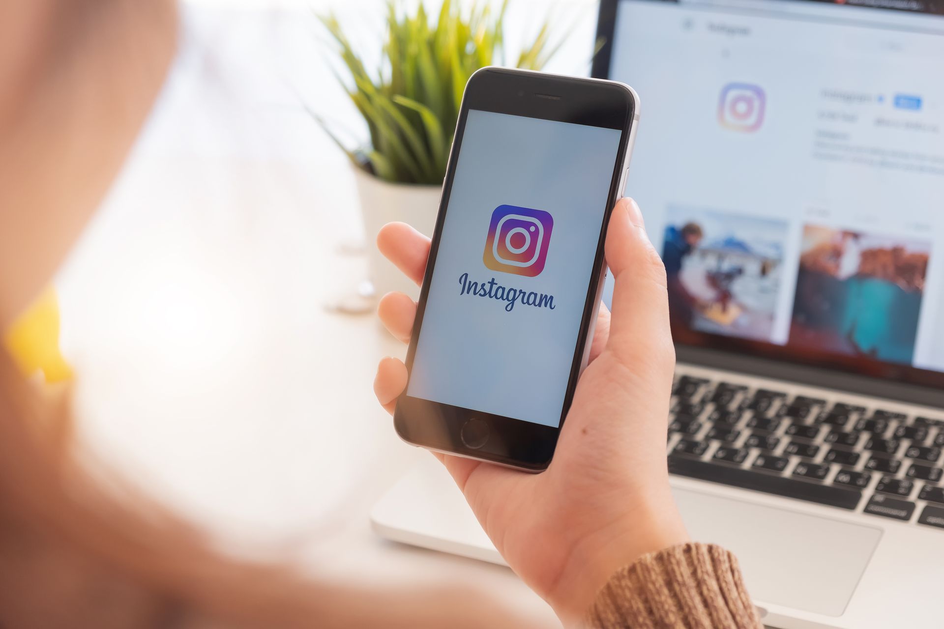 Hand holding a smartphone displaying the Instagram logo beside a laptop on a desk