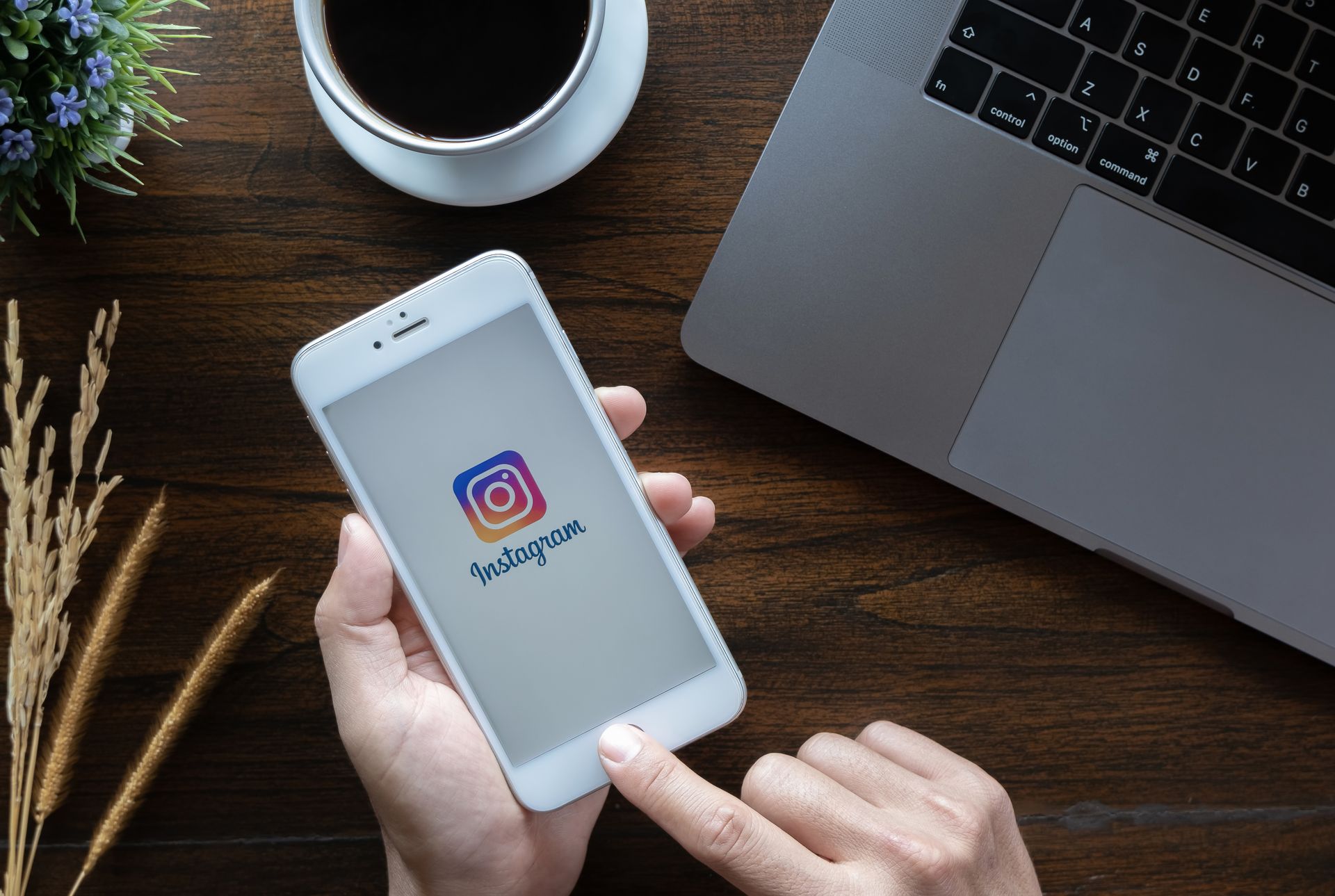 Person holding a phone displaying the Instagram app on a wooden desk with a laptop and coffee cup.