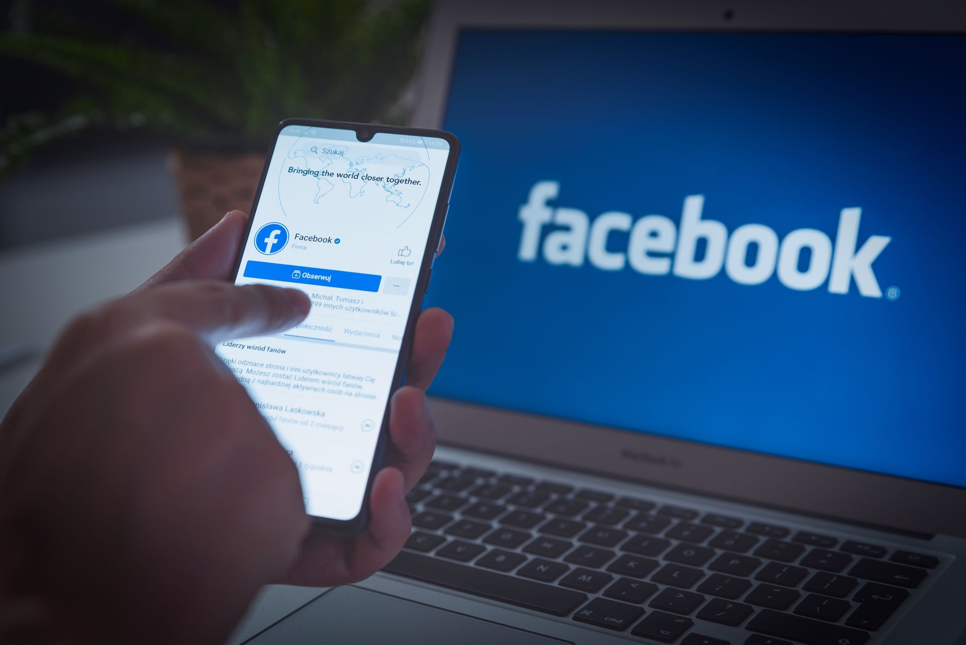 Person using a phone with the Facebook app open, laptop with Facebook logo in background.
