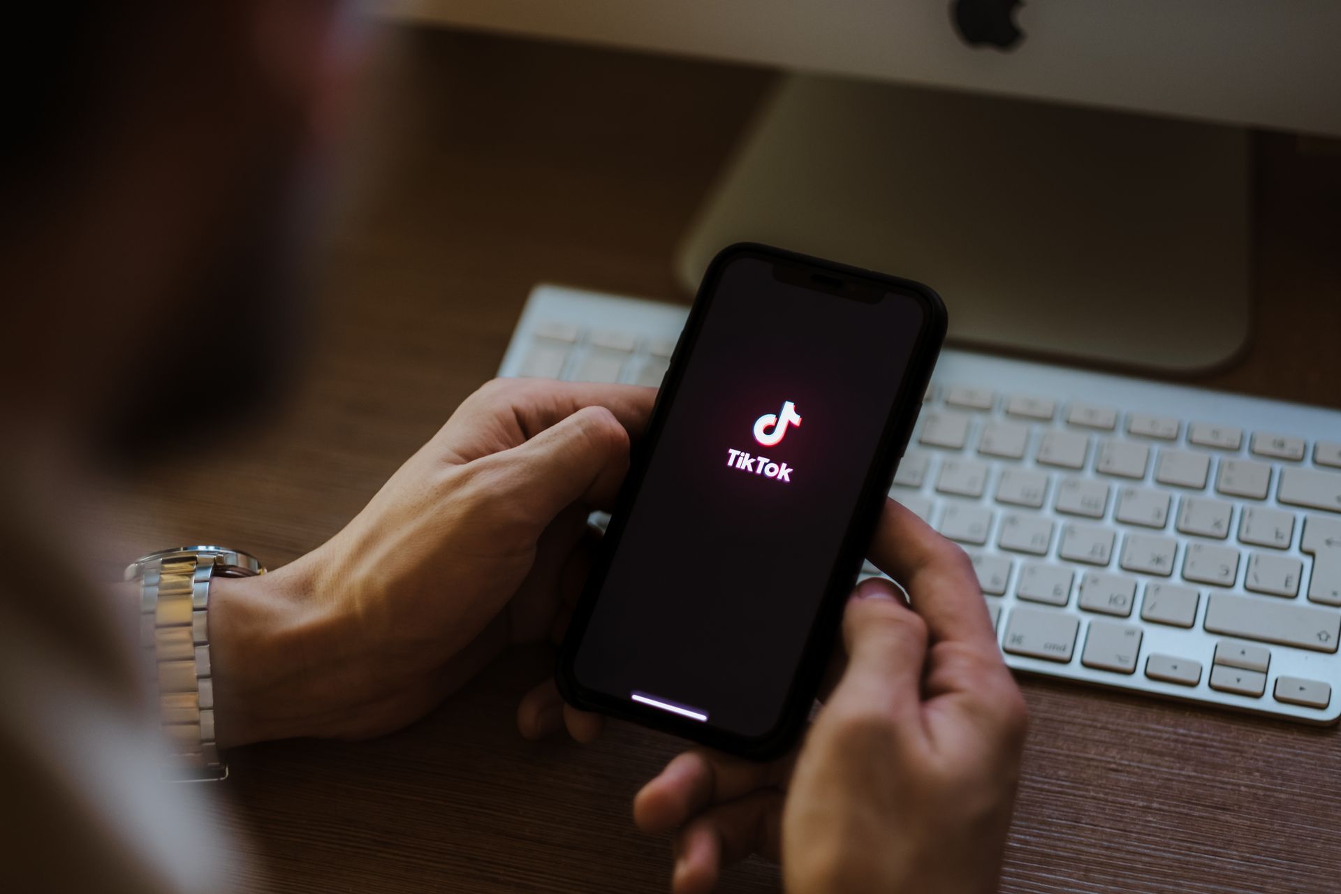 Person holding a phone with the TikTok app open, sitting at a desk with a keyboard and a computer monitor.