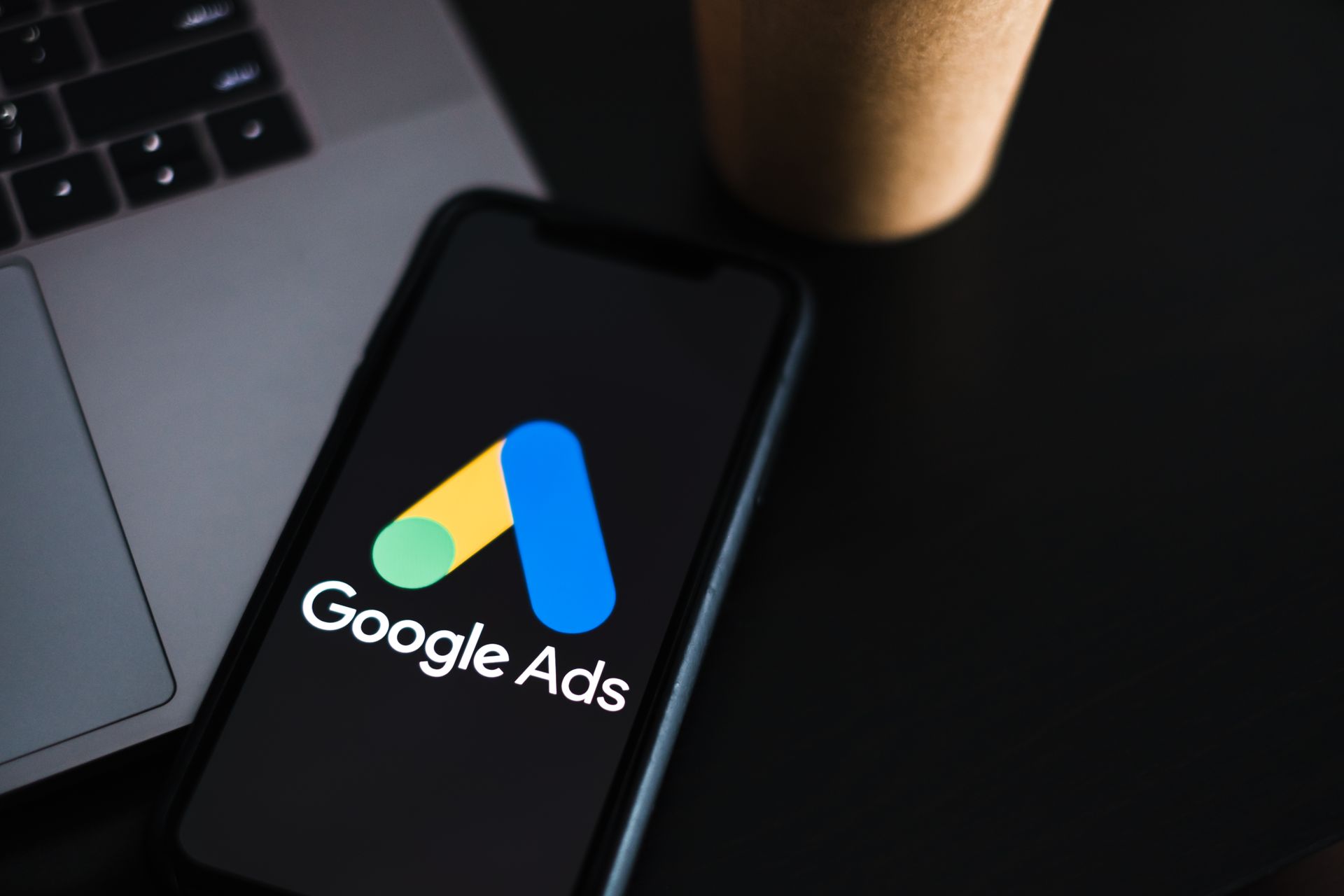 A cell phone with the google ads logo on it is sitting on a table next to a laptop.