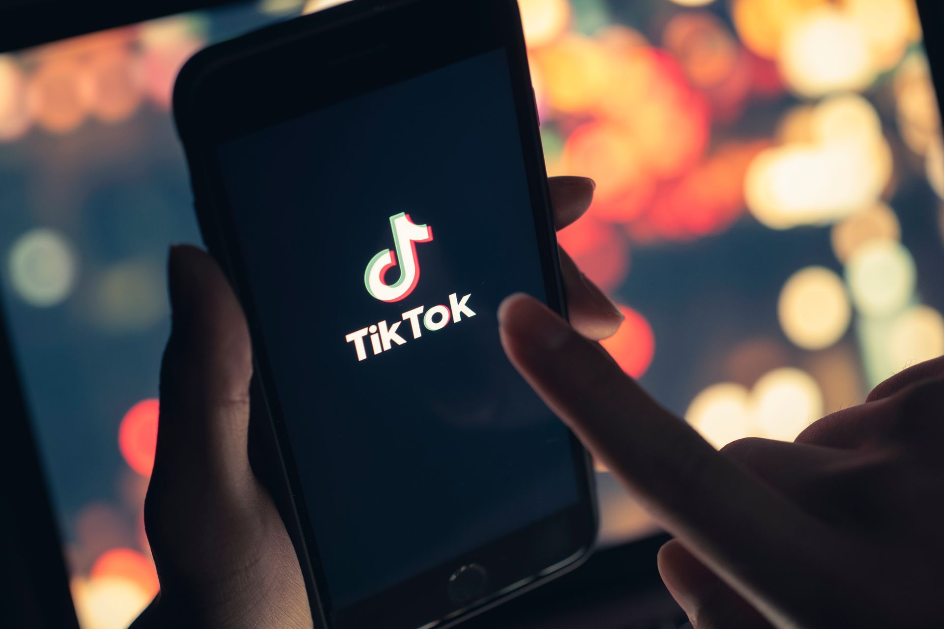 Hand holding a phone displaying the TikTok logo. A finger is touching the screen.