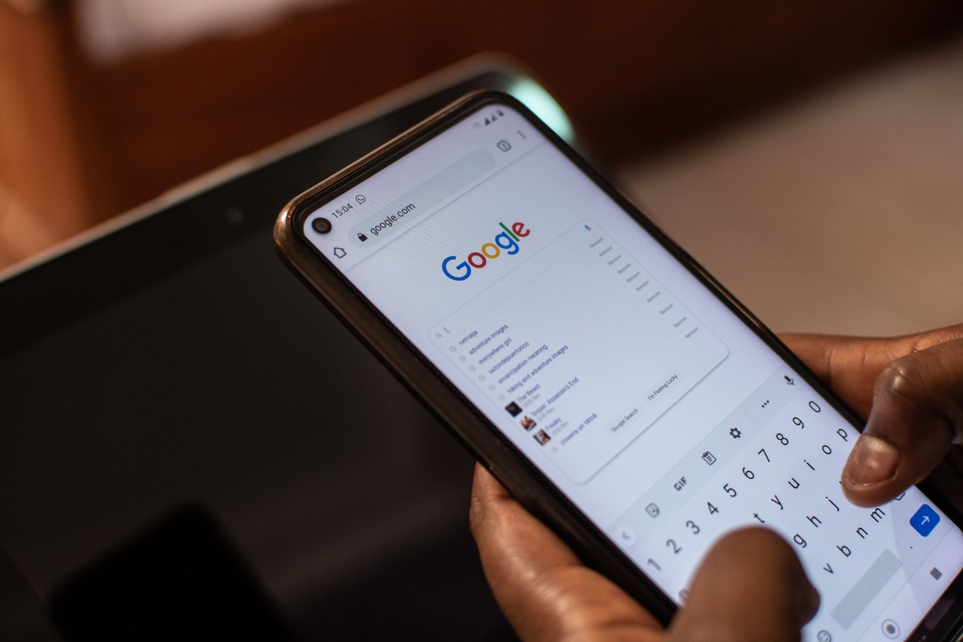 Hands hold a smartphone displaying the Google search homepage with an active virtual keyboard for typing.