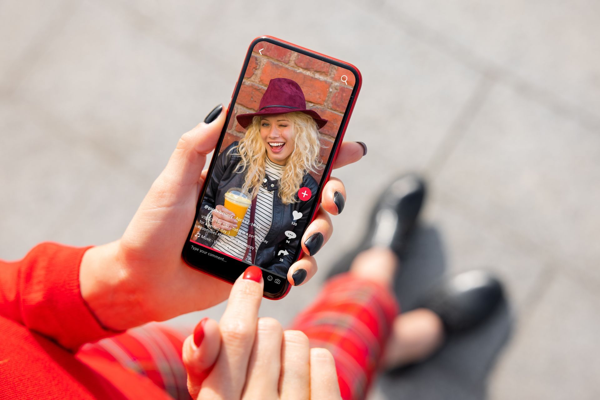 A person holding a red phone with a video of a woman in a hat. Red and black nails. Brick background.