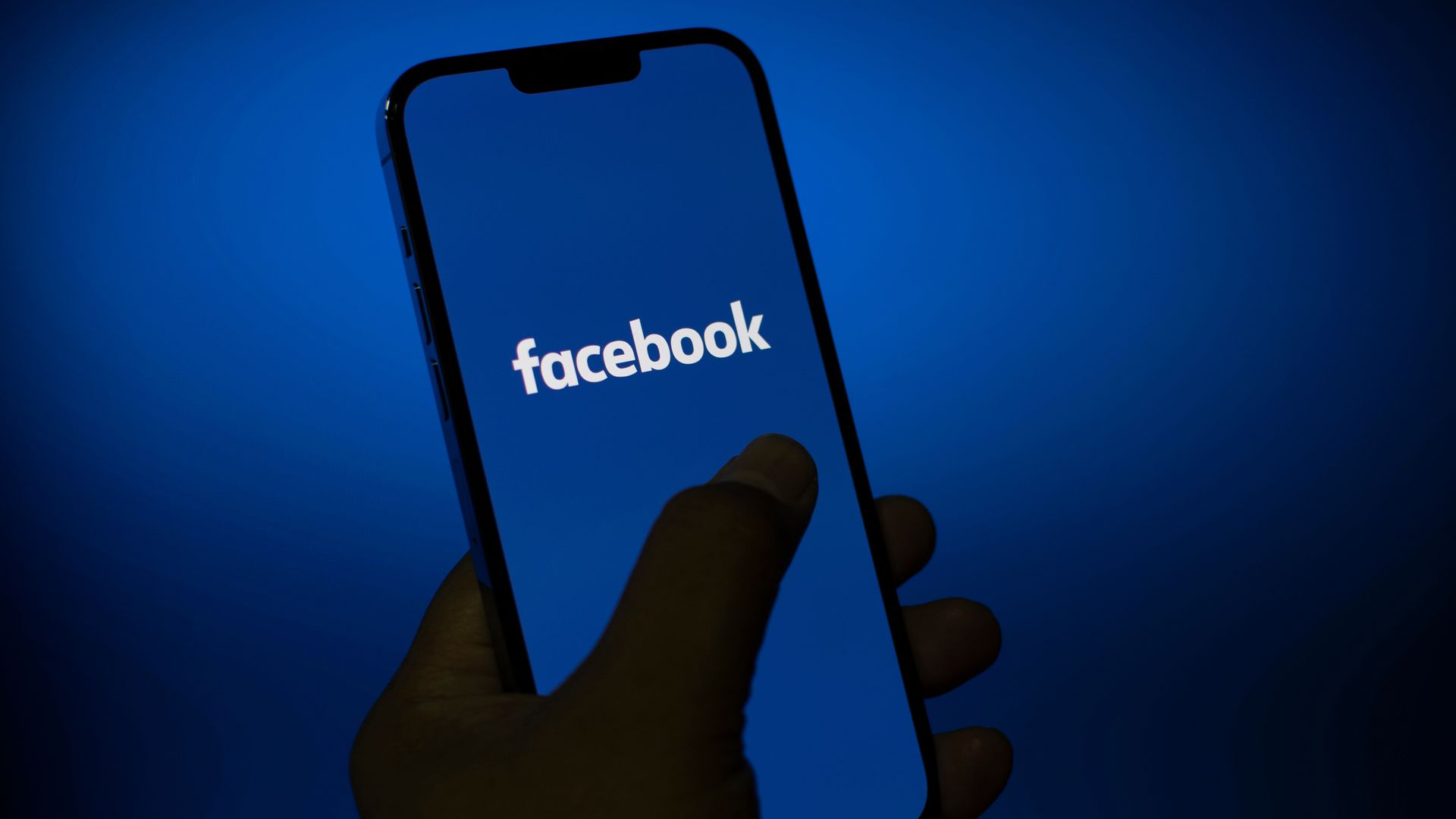 A hand holding a smartphone displaying the Facebook logo on a blue background.
