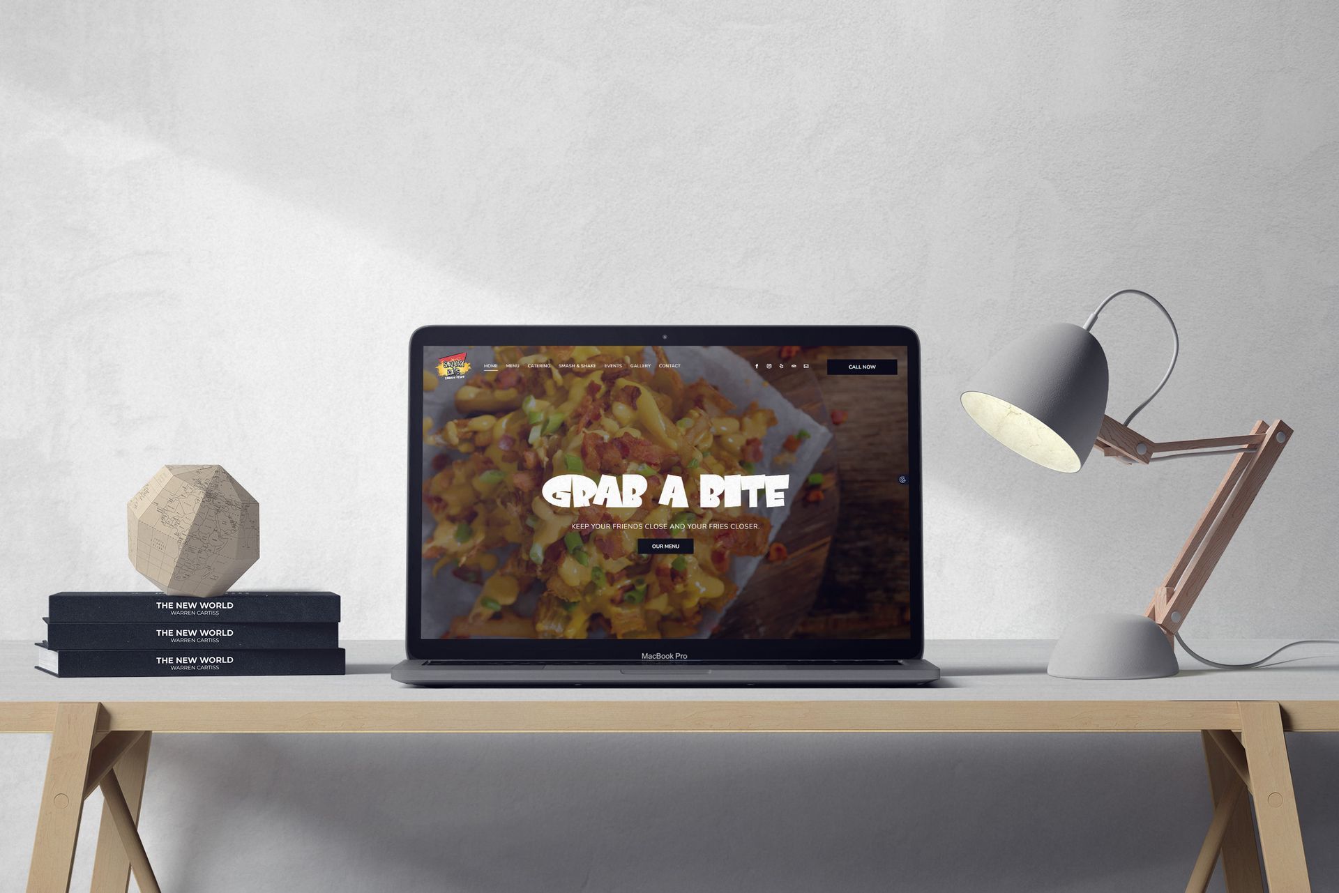 Laptop displaying a website with food, on a desk with a lamp and books.