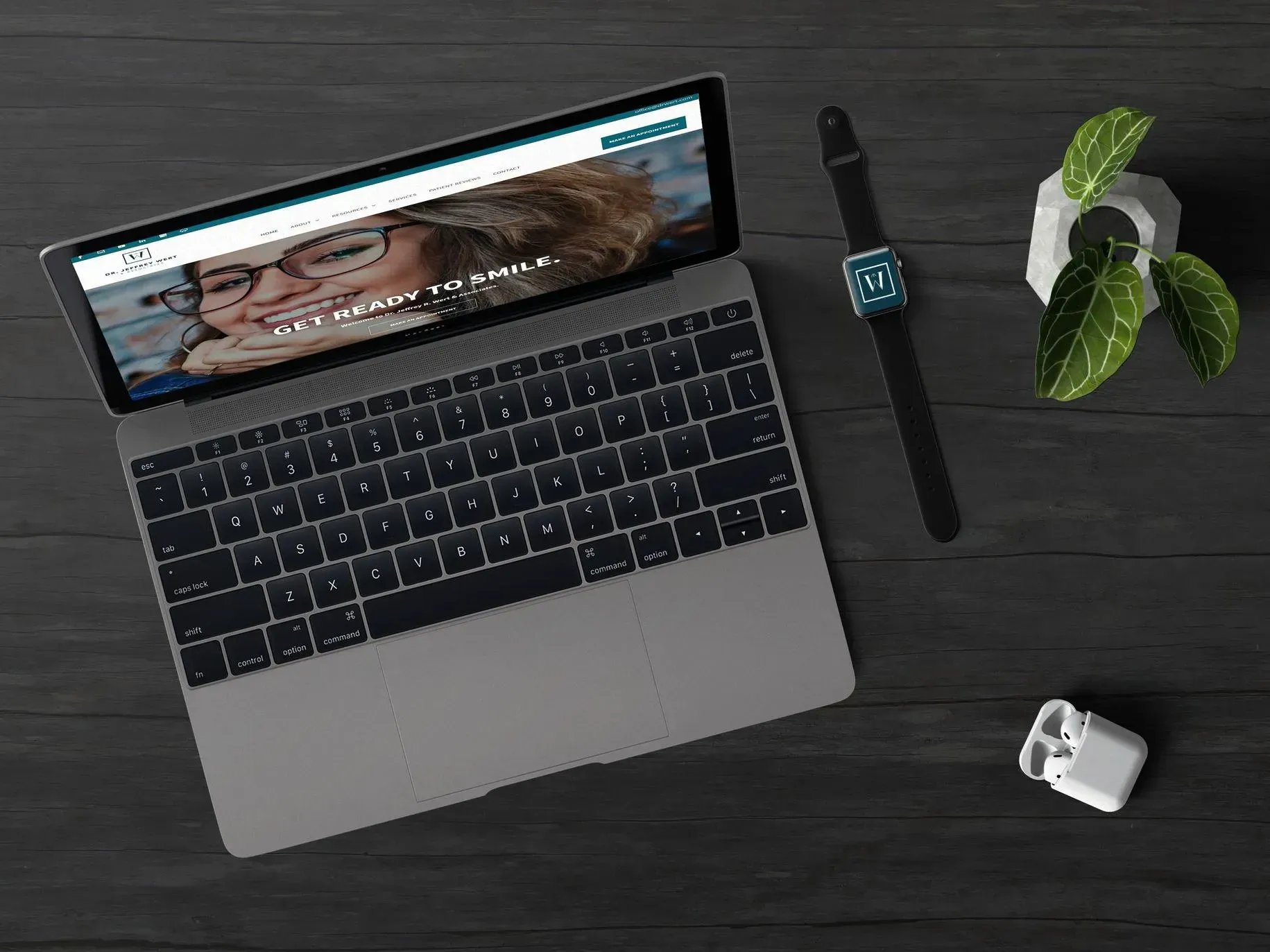 Laptop with website, watch, earbuds, and plant on a dark wooden surface.