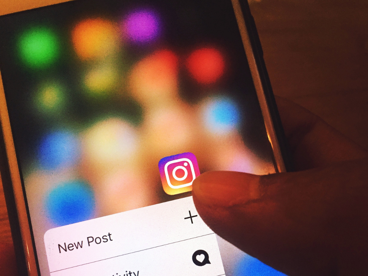 Person's finger touching Instagram app icon on a phone screen, with a blurred background of colorful app icons.