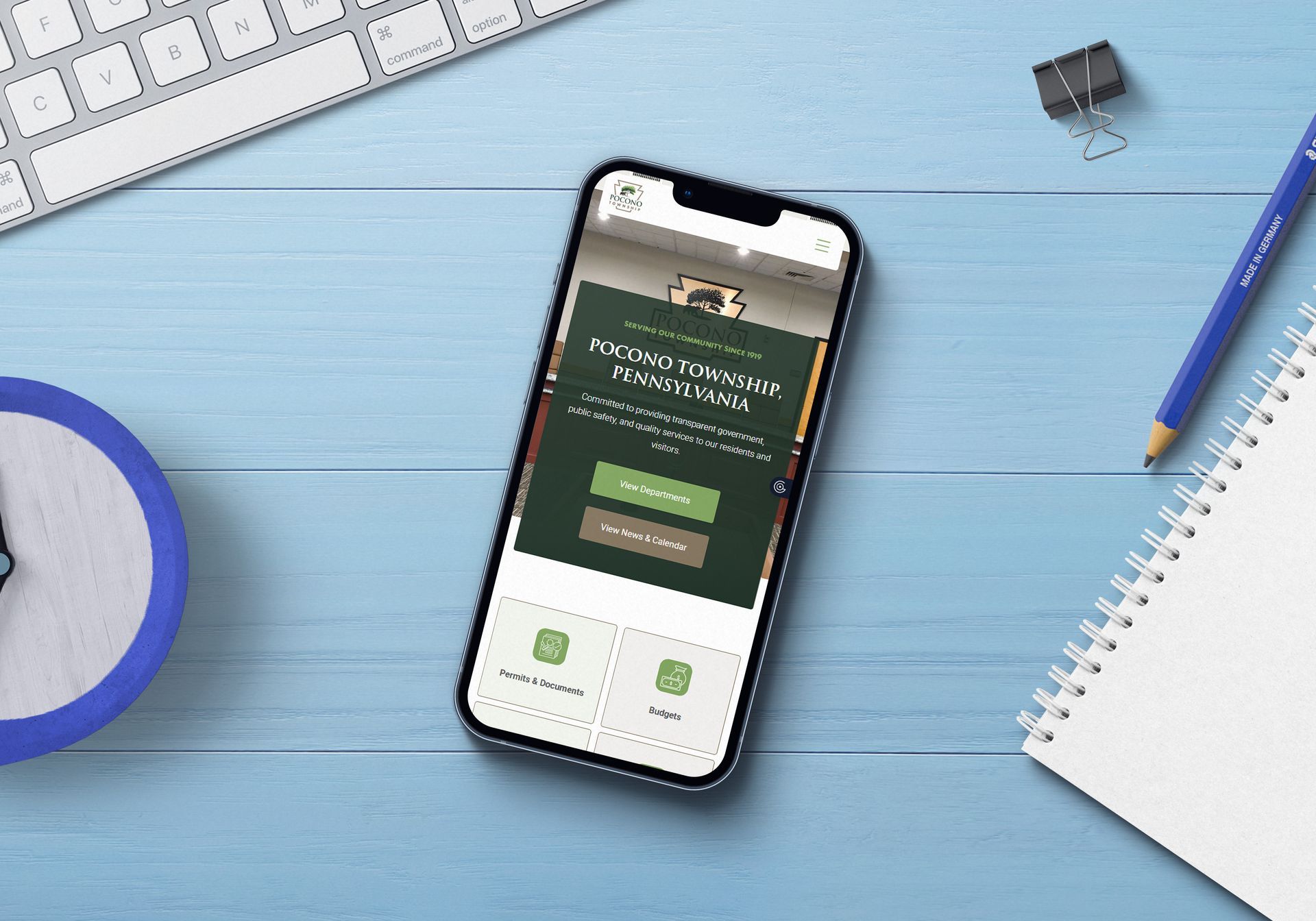 Smartphone displaying website on a blue desk, with keyboard, notepad, clock, and pen.