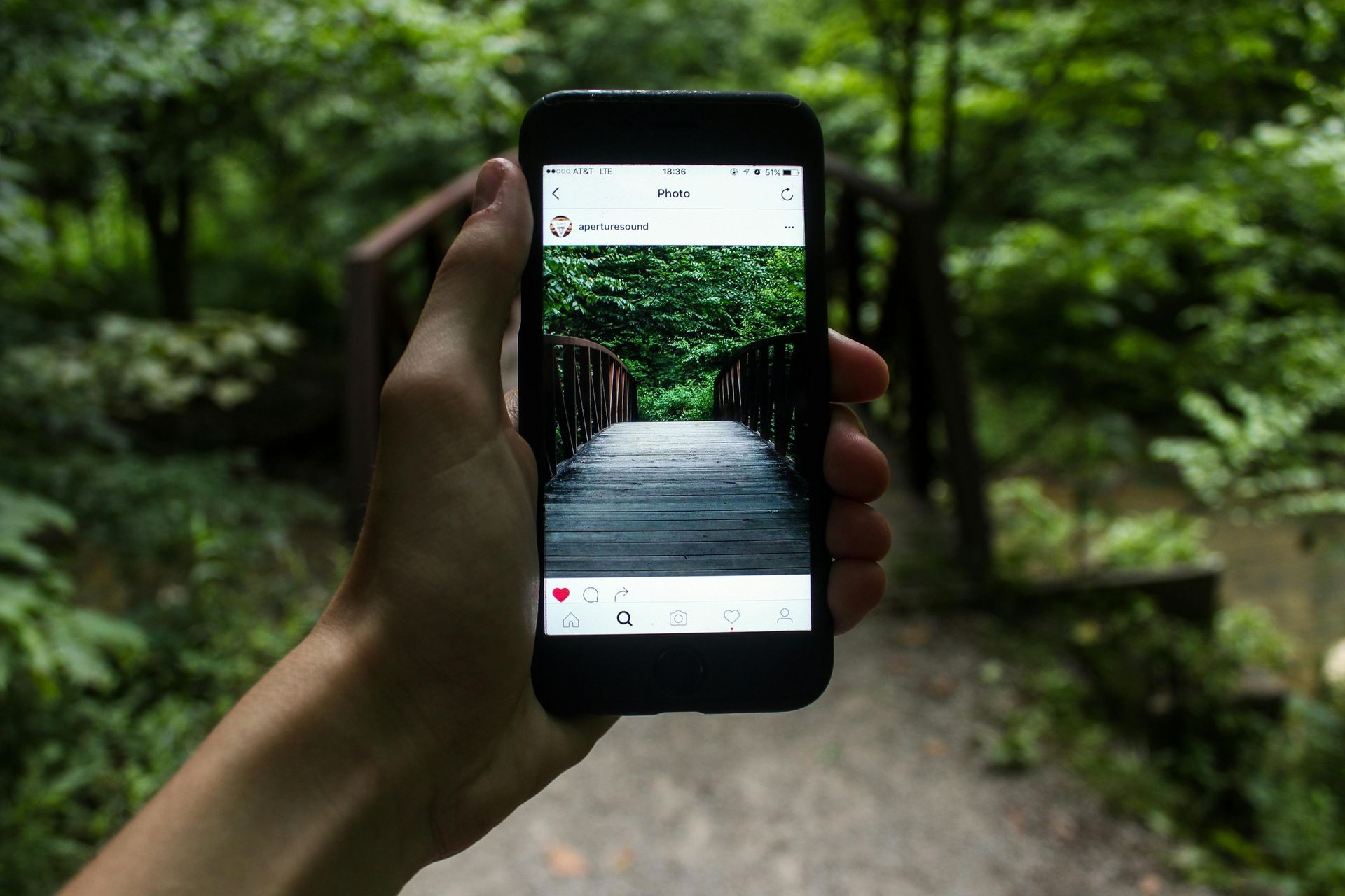 A person is holding a cell phone in their hand and taking a picture of a bridge in the woods.