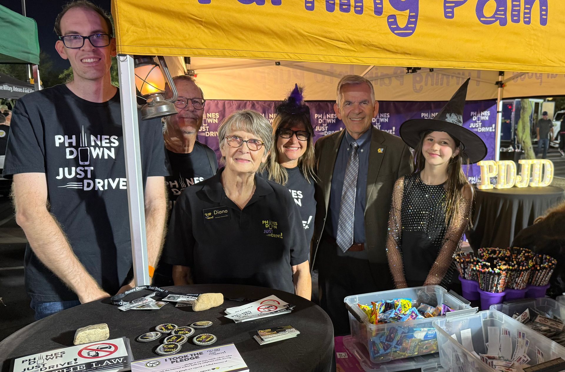 Group of people at a booth with purple banner, various items on display, and a sign that says