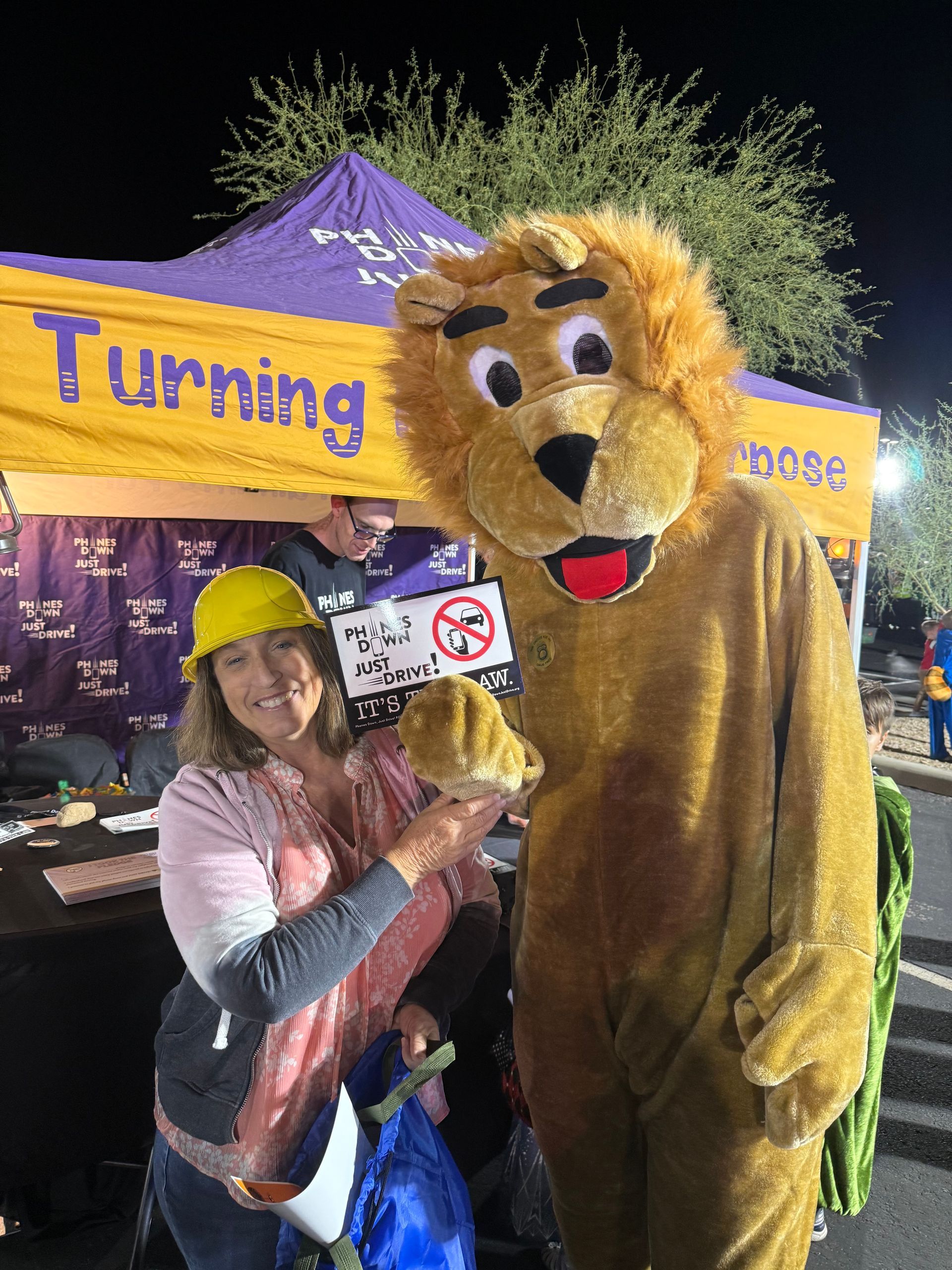 Woman in hard hat holds lion mascot's head at outdoor event with purple tent;