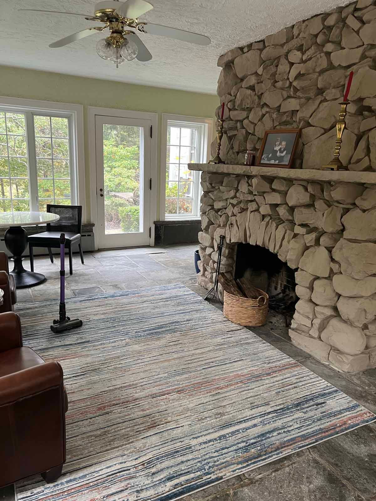Living room with stone fireplace, rug, windows, and ceiling fan.