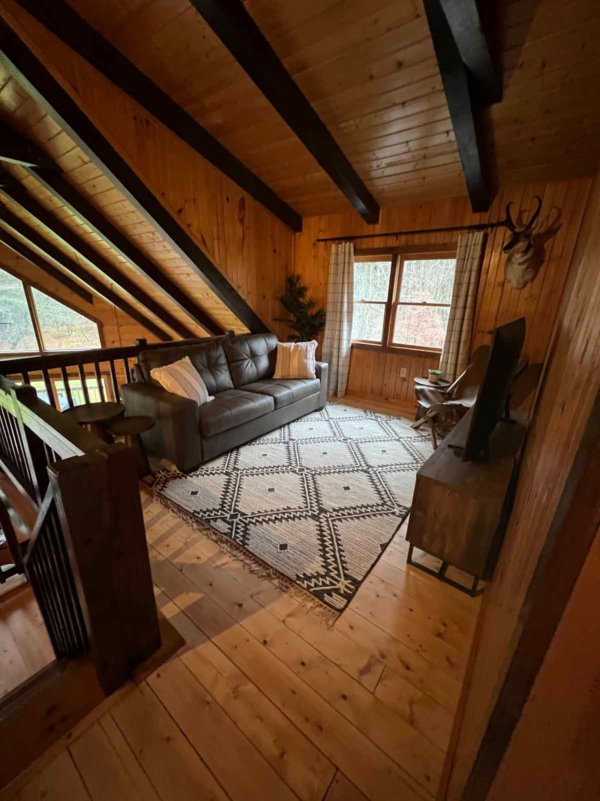 Cozy cabin living room with sofa, rug, TV, wooden beams and walls. Window view, elk head decor.