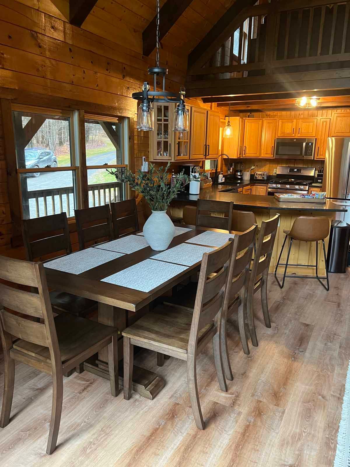 Dining room with large wooden table, chairs, and chandelier. Kitchen in background.