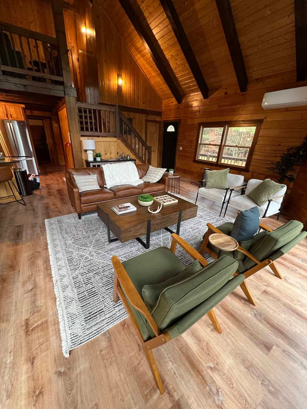 Cozy cabin living room with wooden walls, leather sofa, green chairs, and area rug.