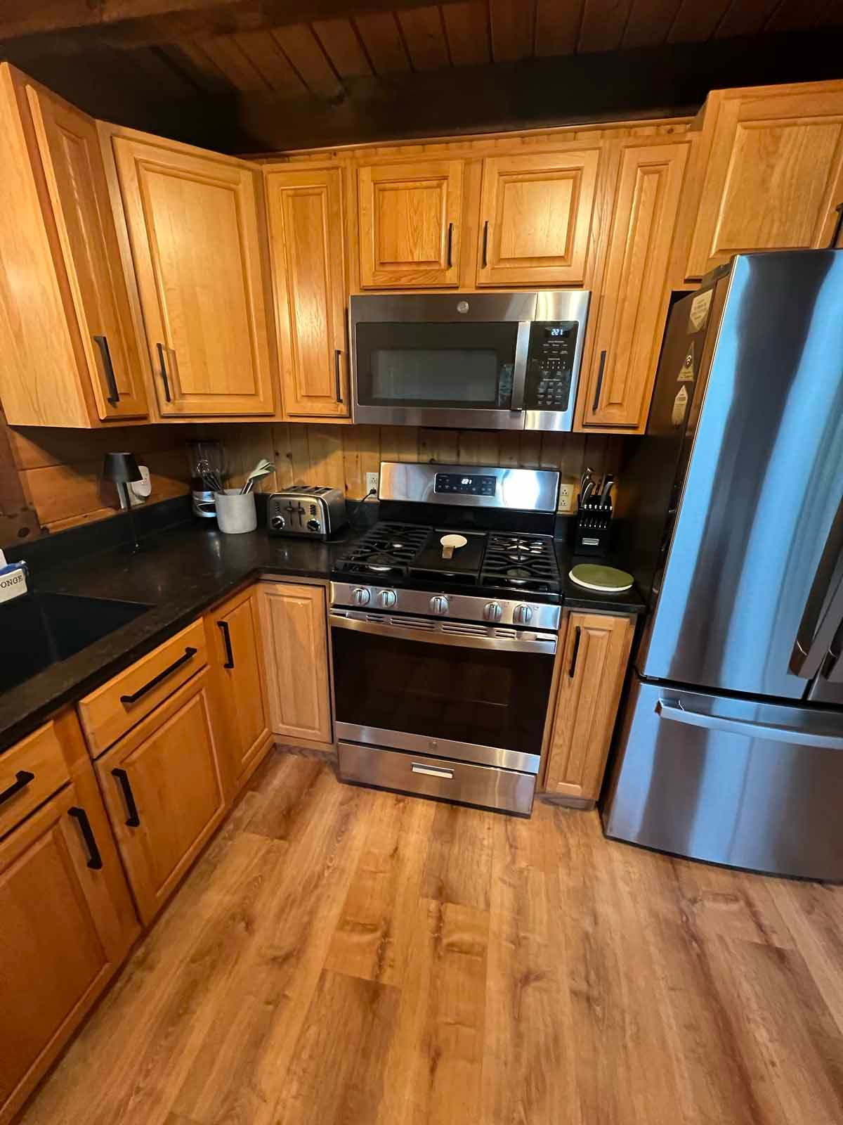 Kitchen with light wood cabinets, stainless steel appliances, black countertops, and wood floors.