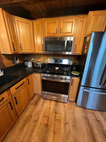Kitchen with light wood cabinets, stainless steel appliances, black countertops, and wood floors.