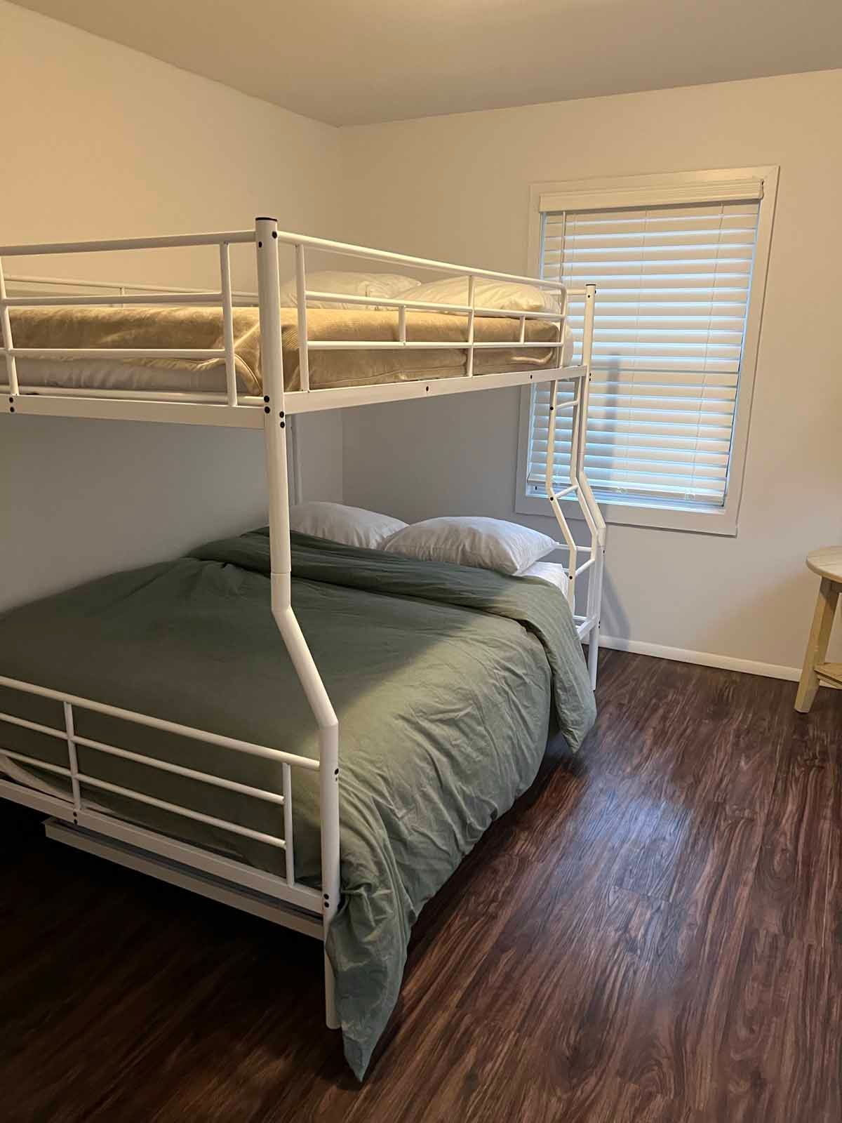 White bunk beds in a room with a window, blinds, and dark wood flooring.