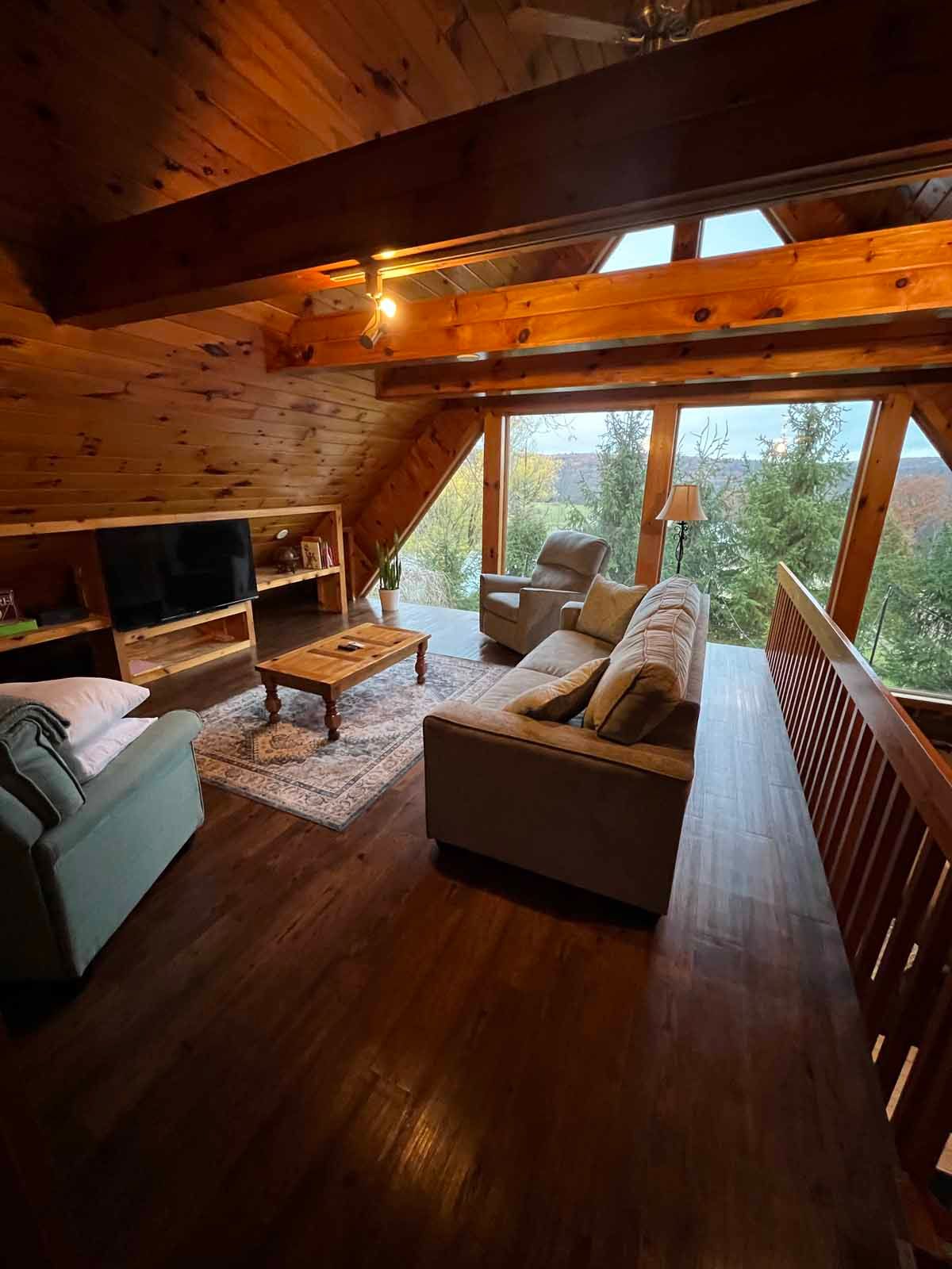 Cozy cabin living room with wood beams, sofas, TV, and large windows with outdoor view.
