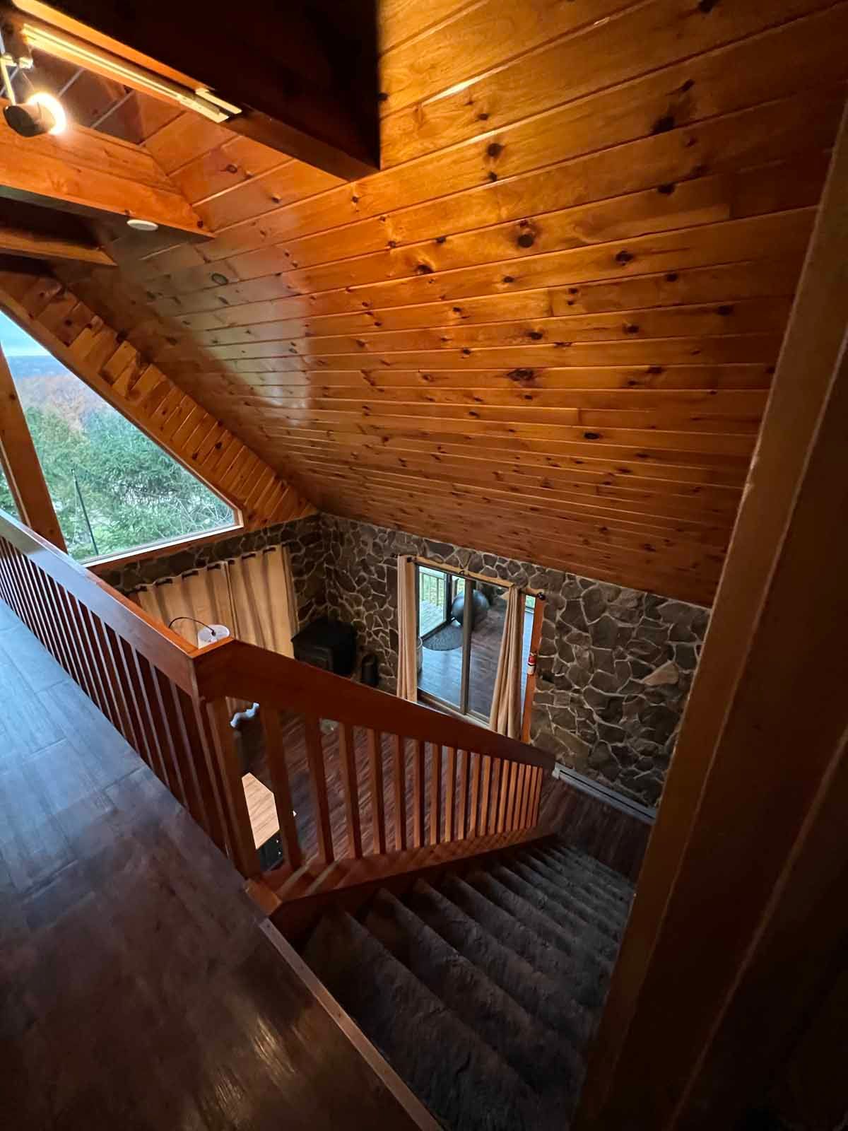 Staircase with wooden railing and brown carpet leading down. Stone wall and wood ceiling above.