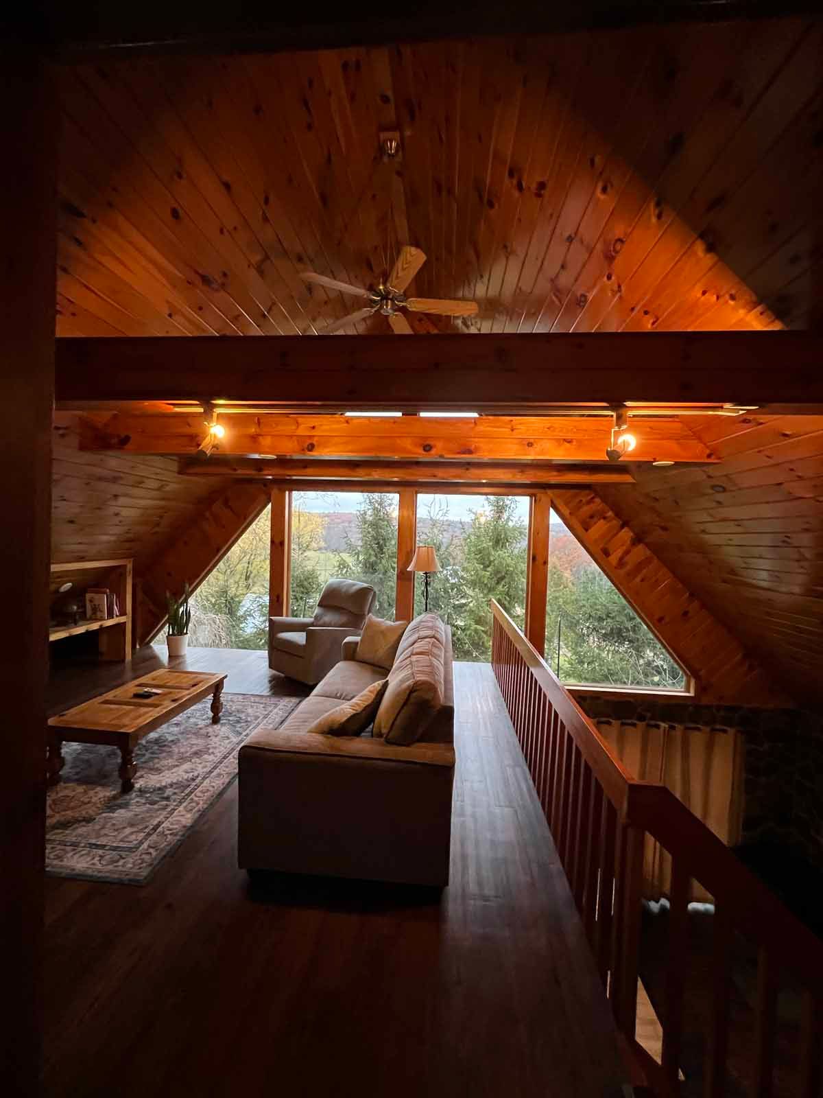 Living room in a cabin with a wooden ceiling and large windows overlooking trees.