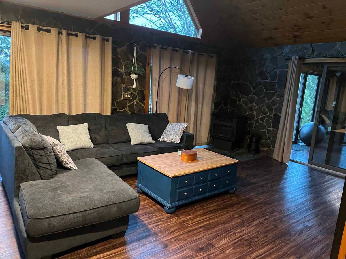 Cozy living room with dark gray sectional sofa, stone wall, coffee table, wood floor, and tan curtains.