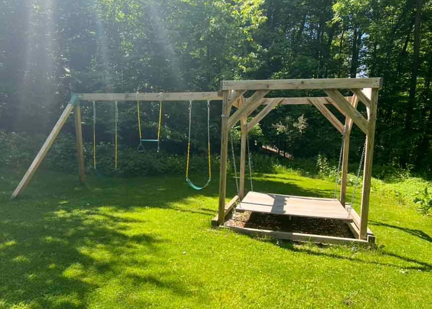 Wooden swing set and playhouse on a grassy lawn with trees in the background.